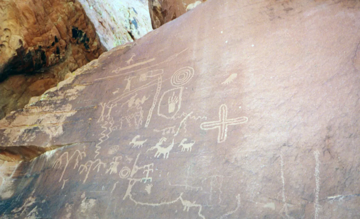 Pale rock art figures of bighorn sheep, people, a spiral and a cross pecked into dark varnish on a red sandstone face